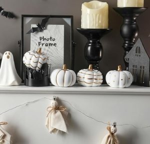 White and gold ceramic pumpkins sit on a mantelpiece along with a string of paper ghosts, candles and a picture frame