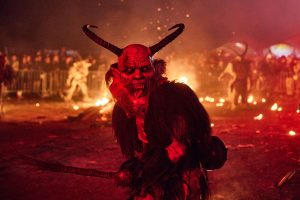 Krampus stands in front of fire in the street with his tongue wagging.