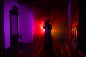 A witch stands inside a house with creepy lighting, holding a pitchfork or a broom